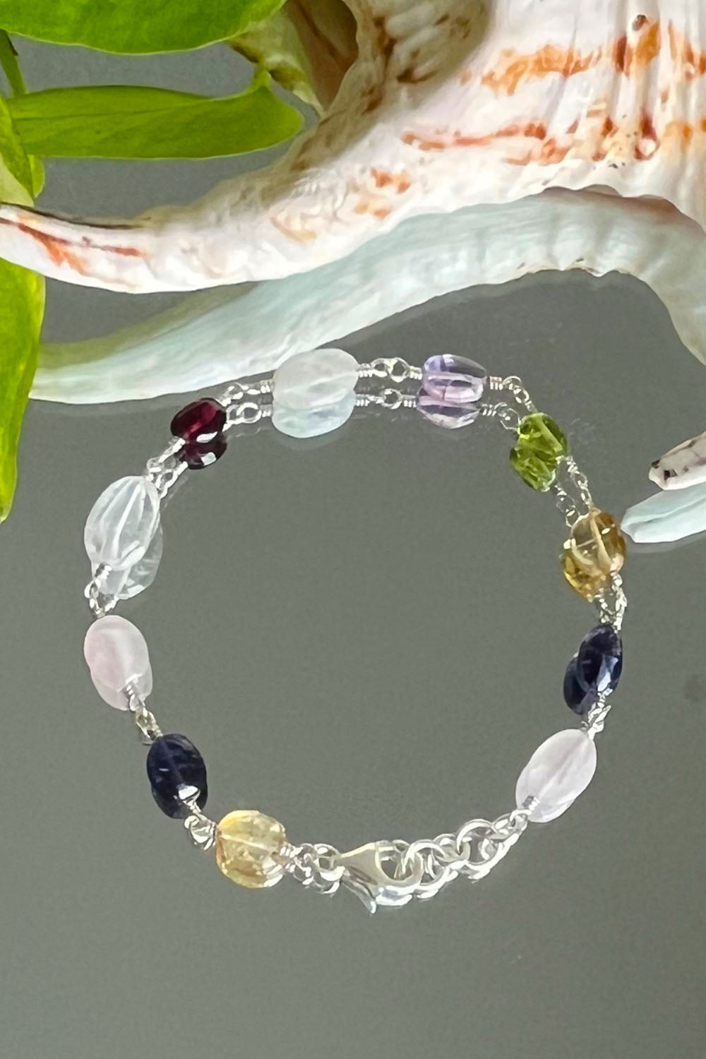 Astra multigemstone silver bracelet beside a shell and green leaves